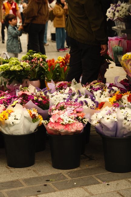 A variety of fresh floral bouquets displayed for flower delivery and gifting on a market stall, arranged in black buckets on a paved surface. The arrangements include pink, white, purple, and yellow f