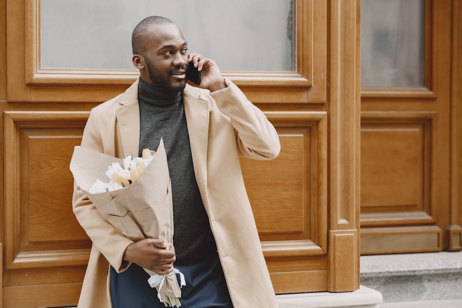 A man with dark skin, a neatly trimmed beard, and a friendly expression, is standing outdoors against a wooden-paneled wall with large glass windows. He is dressed in a beige overcoat over a dark grey