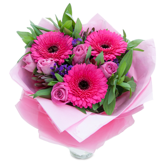 A beautifully balanced spring bouquet where three fuchsia-pink gerbera daisies command attention with their open, sunburst petals and dark centres that provide striking contrast. Around these, blush-to-lavender roses sit in close buds, their velvety layers curling inward and offering a softer, romantic note. Interspersed are sprigs of tiny purple statice that float like confetti between the larger blooms, bringing delicate structure and a hint of wildflower charm. Generous, leafy greenery-broad, dewy leaves reminiscent of salal or eucalyptus-wraps the arrangement in a green embrace, giving weight and a fresh garden scent that complements the flowers' fragrance. The bouquet is dressed in a pale matte pink tissue inner wrap and an outer veil of sheer, crinkled fuchsia-pink cellophane that catches light and intensifies the colour story; the florist's folds reveal the top rim of a clear glass vase, indicating the stems are ready to sit in water and be enjoyed immediately. In soft daylight the petals take on a luminous quality and the combination of textures-from the smooth rose petals to the papery statice-feels crafted and comforting. This arrangement suits many moments: a warm anniversary gift for a couple on Stockwell Park Walk, a birthday gesture to brighten a flat near Clapham Common, or simply a thoughtful piece to bring cheer into a local home. It reads as thoughtful, joyful, and distinctly hand-touched by an attentive florist.